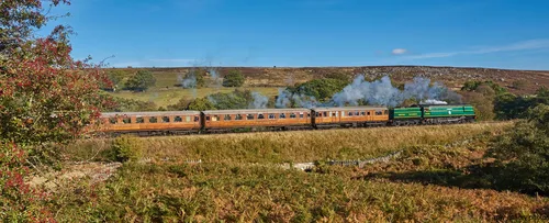 This landscape photograph captures a steam train traveling through the rural scenery of the North Yorkshire Moors in the United Kingdom during an autumn afternoon in 2018. The North Yorks steam train, featuring traditional carriages and a locomotive emitting visible steam, is depicted as it moves across the open countryside, with natural moorland and rolling hills in the background. The image clearly represents transport in North Yorkshire, showcasing the historical significance of train travel in this region.
